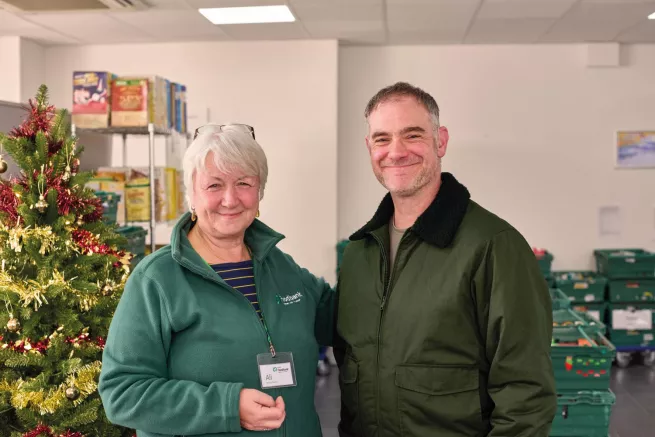 A food bank volunteer stands next to a visitor, both smiling into the camera