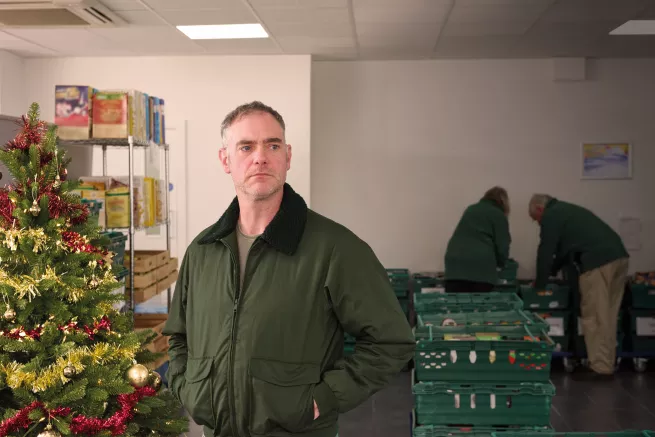 A man standing next to a Christmas tree at a food bank. In the background two people are handling crates of donated food.