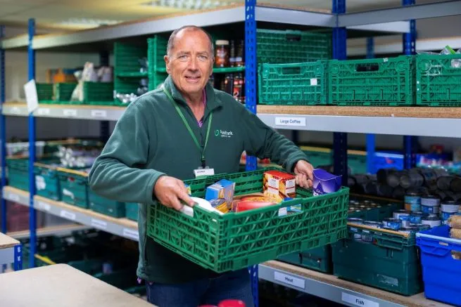 A man volunteering in a food bank warehouse, holding a crate of food