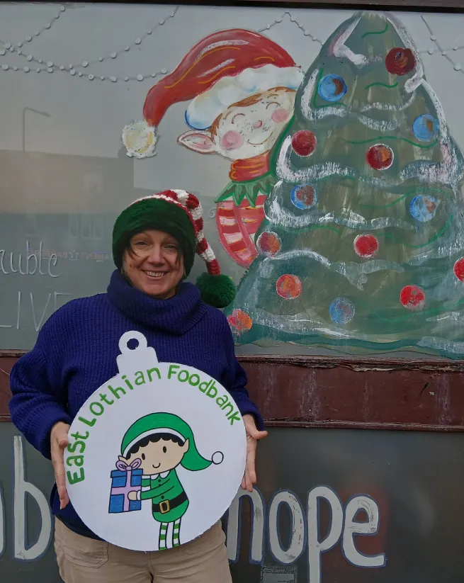 Person in an elf hat standing in front of a Christmas decorated window holding a carboard bauble with an elf drawn on it