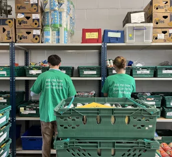 Two people stood in a warehouse full of green creates containing food items. The people have their backs to the camera and are sorting food items.