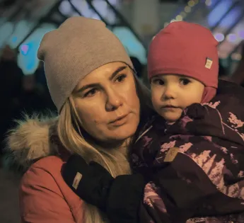 Woman holds a young child, who looks into the camera. They are outside wearing winter clothes.