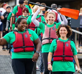 Three participants of a dragonboat race celebrate. All are smiling towards the camera and one raises a paddle into the air.