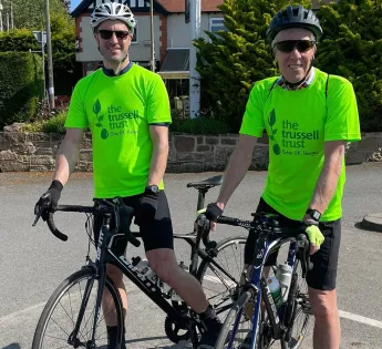 Two fundraisers smile into the camera as they stand with their bikes. Both are wearing bright green Trussell Trust t-shirts, safety helmets and sunglasses.