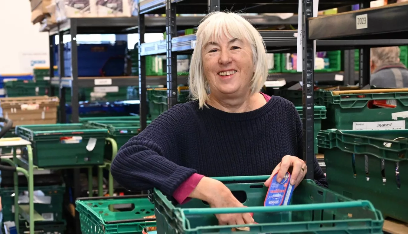 Smiling woman holding food items behind stack of green crates in food bank warehouse. 