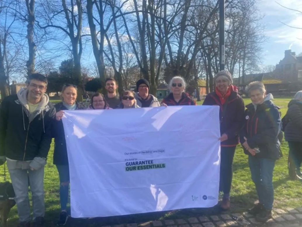 A group of 9 campaigners stood outside on a sunny day with a tablecloth reading 'Stories of Hope and Hardship. Guarantee Our Essentials.'