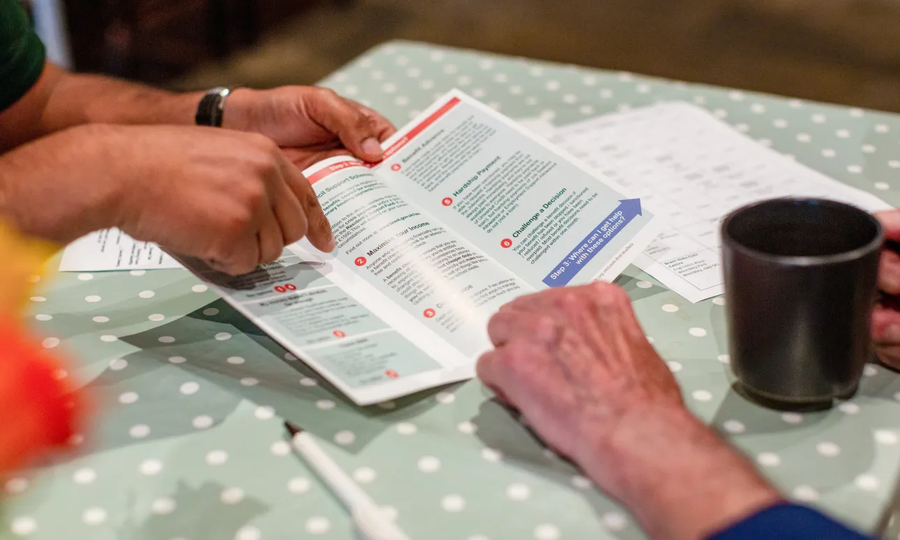 A detail of information leaflets being discussed at a table at the food Bank, with hands and a cup of tea visible.