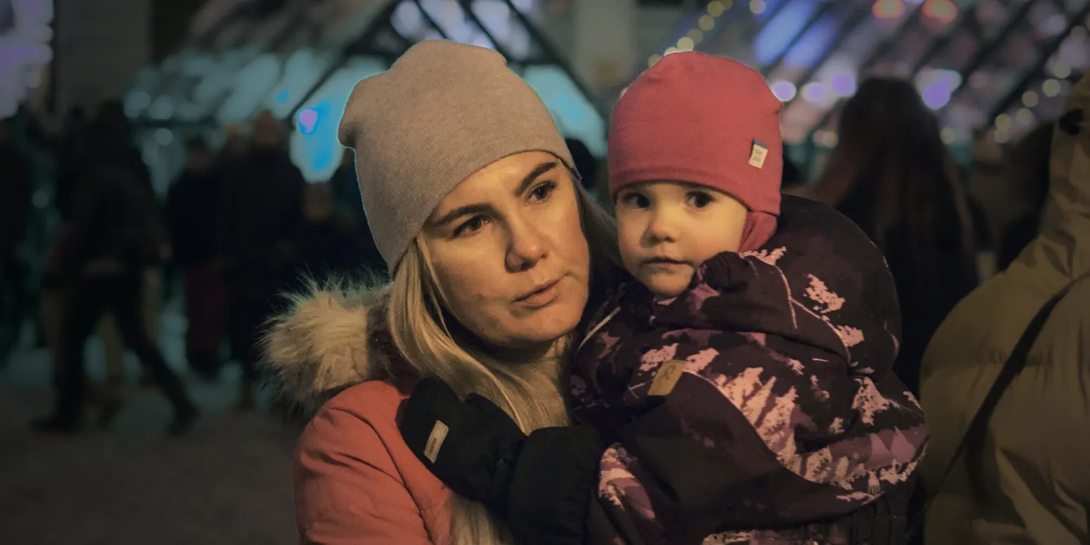 Woman holds a young child, who looks into the camera. They are outside wearing winter clothes.