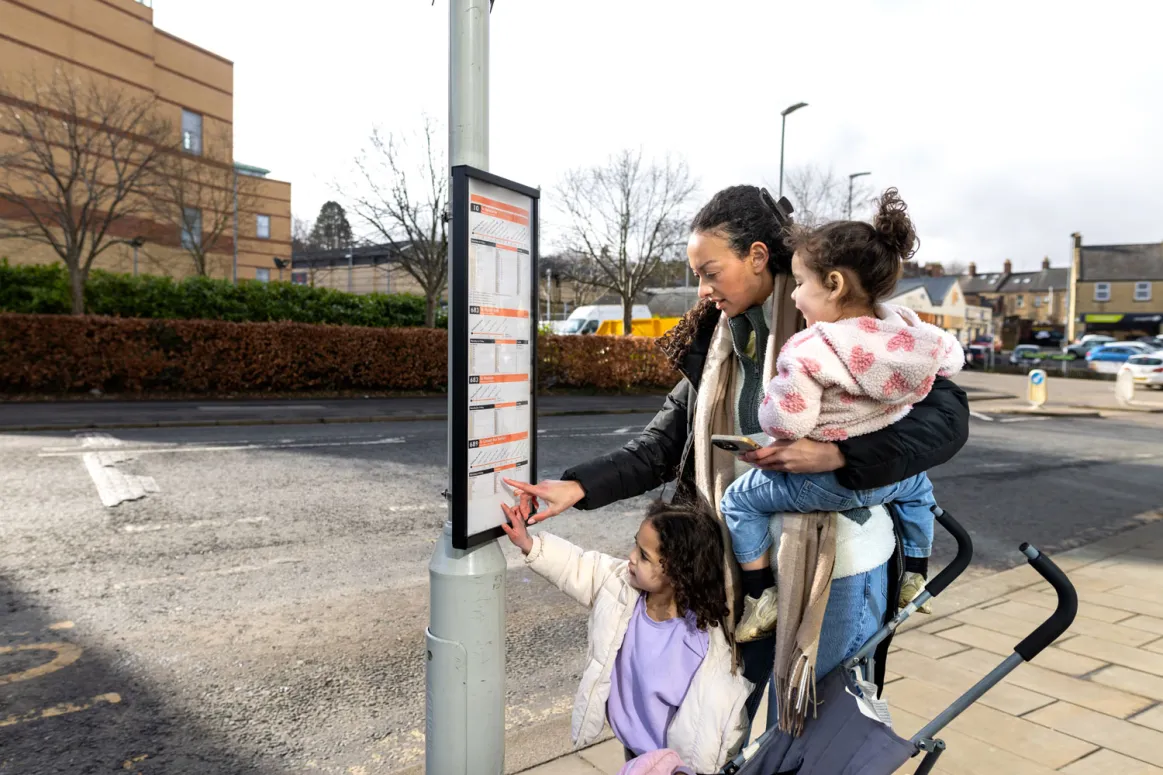 A woman and her two children at a bus stop, looking at the bus timetable