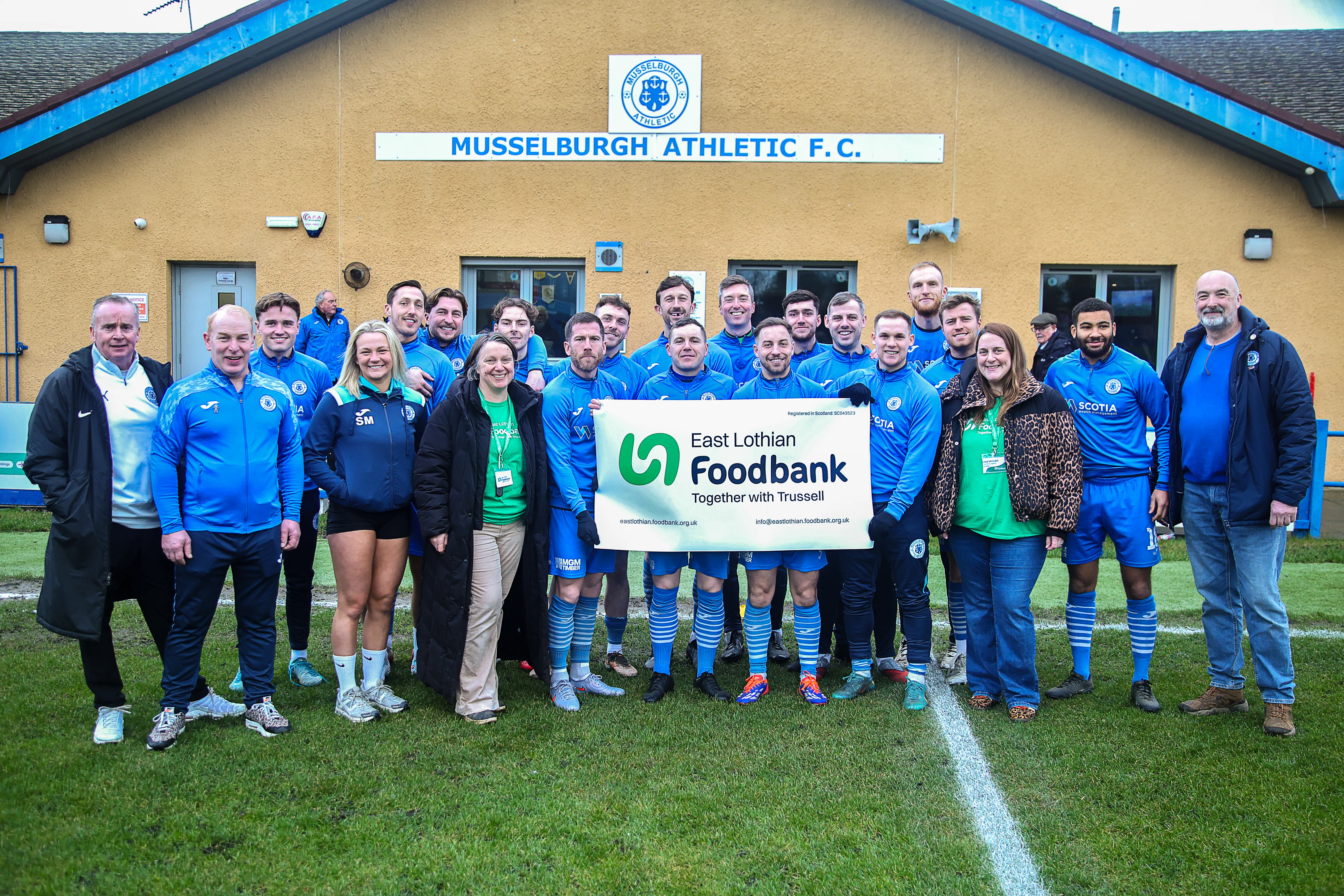 Musselburgh Athletic FC team standing with some of East Lothian Foodbank team hold and East Lothian Foodbank banner