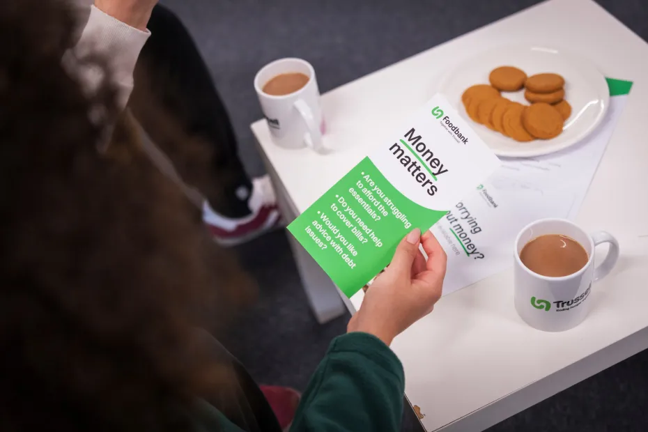 The hands of a food bank volunteer with a money matters leaflet, at a table with with cups of tea and a plate of biscuits.