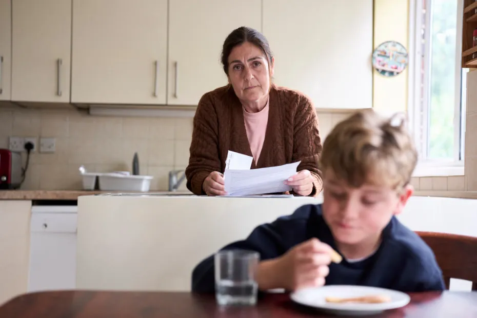 Woman in kitchen with bills, child sitting at table with a snack