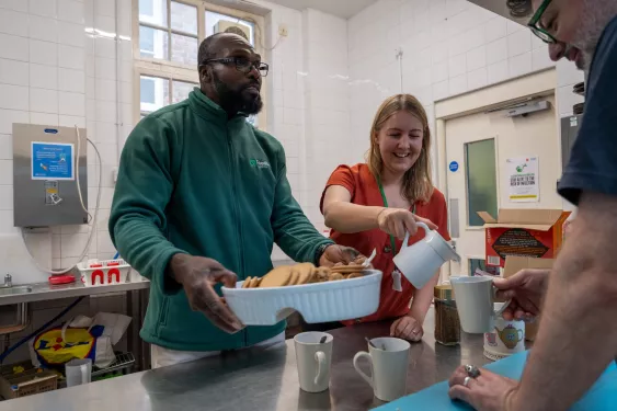 A man and a woman at Lewisham food bank making tea and offering a bowl of biscuits.