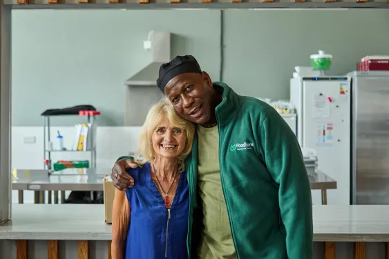 A man and a woman embrace in a food bank kitchen. He is wearing a cook's cap and a green Trussell fleece, while she is wearing a blue blouse.