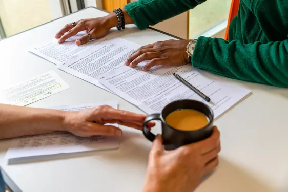Two people at white table, one drinking a cup of tea and the other filling out paper forms