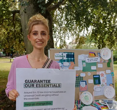 Woman stands next to a colourful campaign stall holding a poster reading 'Guarantee Our Essentials.'