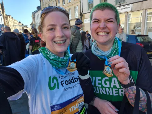 A pair of female runners, with one wearnig a Cirencester Foodbank running shirt proudly raising their medals which hang around their necks