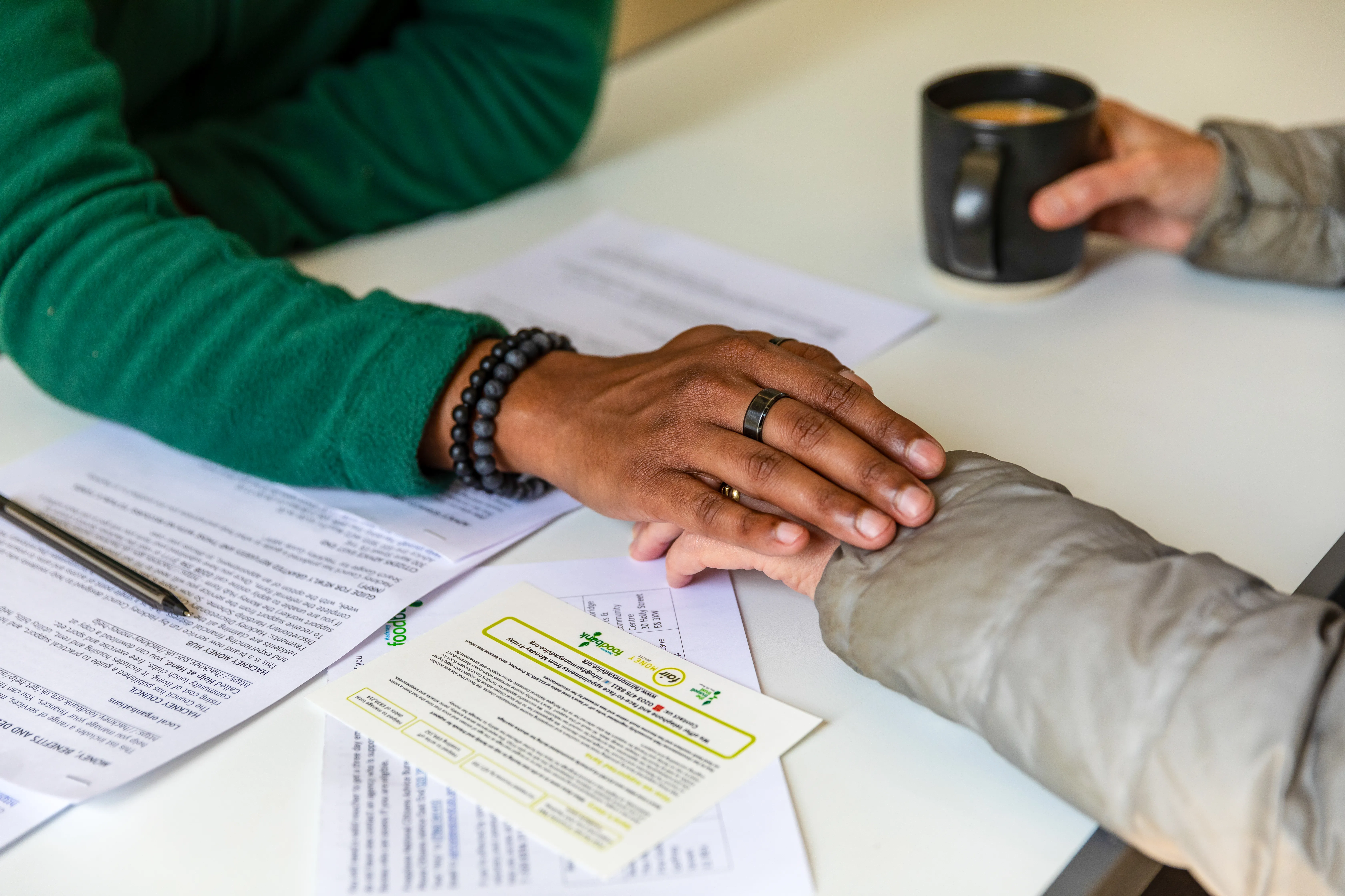 A picture of two people, but only their forearms and hands are showing, resting on a table, outstretched towards each other, with one persons hand on top of the other. A cup of tea is held by the person whose hand is being held.