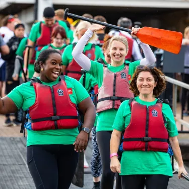 Three participants of a dragonboat race celebrate. All are smiling towards the camera and one raises a paddle into the air.