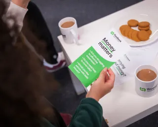 The hands of a food bank volunteer with a money matters leaflet, at a table with with cups of tea and a plate of biscuits.