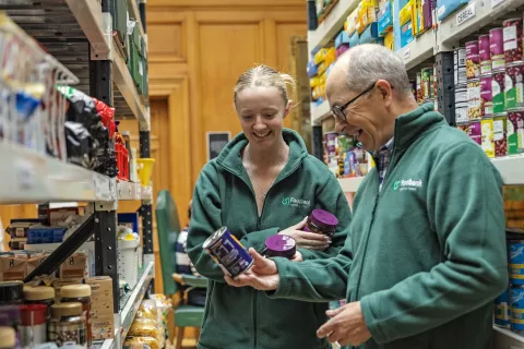 Two members of staff smiling whilst looking at food supplies in the aisles of the Peeblesshire food bank