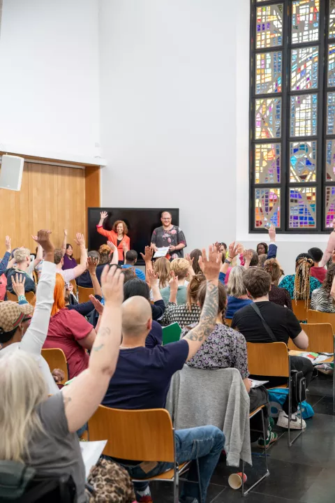 A large gathering of people inside a church room at a Legislative theatre event including lived experience partners and audiences. A people are stood up at the front of the room and the audience are sitting away the camera with the hands raised up.