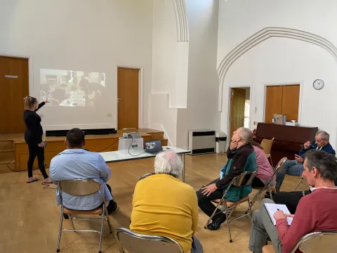 Group of people sat in a large hall facing a screen. A woman gestures towards the image on screen as if explaning it.